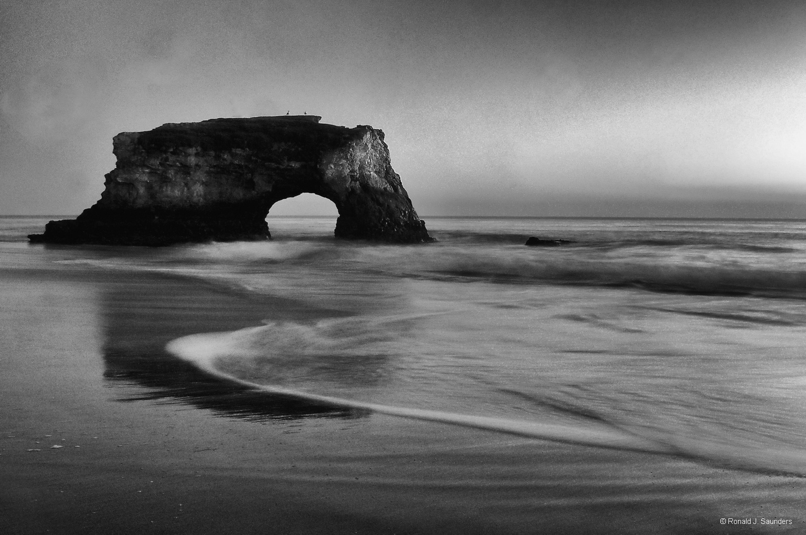 A sunset view of the arch at Natural Bridges State Beach. The beach features picturesque arches cut out by ocean waves in a sandstone...