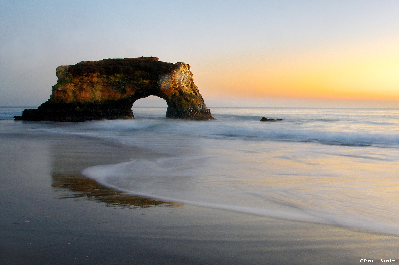 A sunset view of the arch at Natural Bridges State Beach. The beach features picturesque arches cut out by ocean waves in a sandstone...